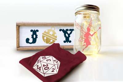 A canning jar, wood framed sign, and shirt are arranged on a white background. The jar has fairy lights inside and is decorated with a fairy silhouette. The shirt has a 20-sided die. The sign says "JOY" with a bell for the O.