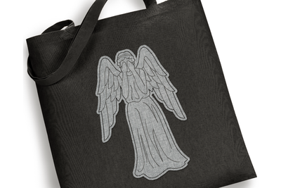 A black tote bag sits on a white background. Embroidered onto the bag is the image of an angel statue hiding its face, done in redwork/linework style. The angel has raggy applique fabric behind the design in a grey fabric.