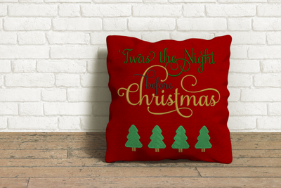 A red throw pillow sits on a wooden floor against a white brick wall. Embroidered onto the pillow in shades of gold, green, and black is the phrase "Twas the night before Christmas" with a row of 4 Christmas trees beneath.