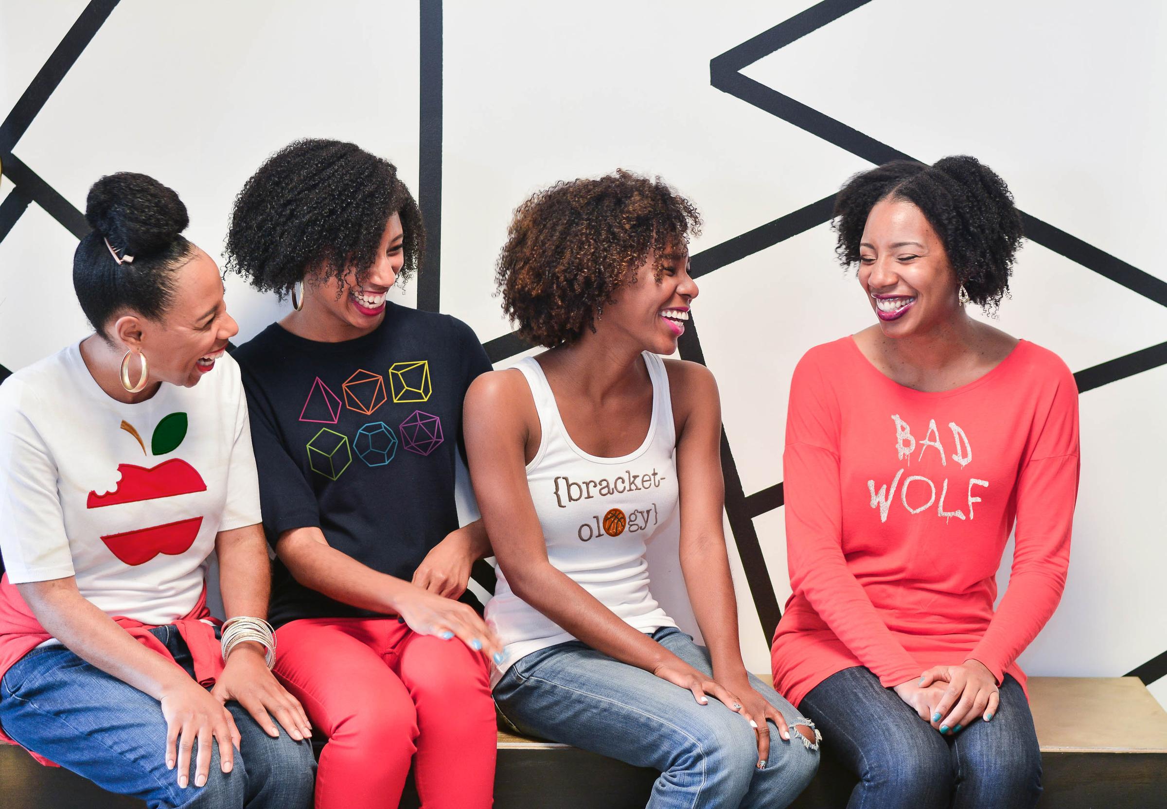 Four black women are sitting on a bench in front of a white wall with geometric black artwork. The women are smiling and laughing. Each woman has a shirt with an applique or embroidery design on the front.