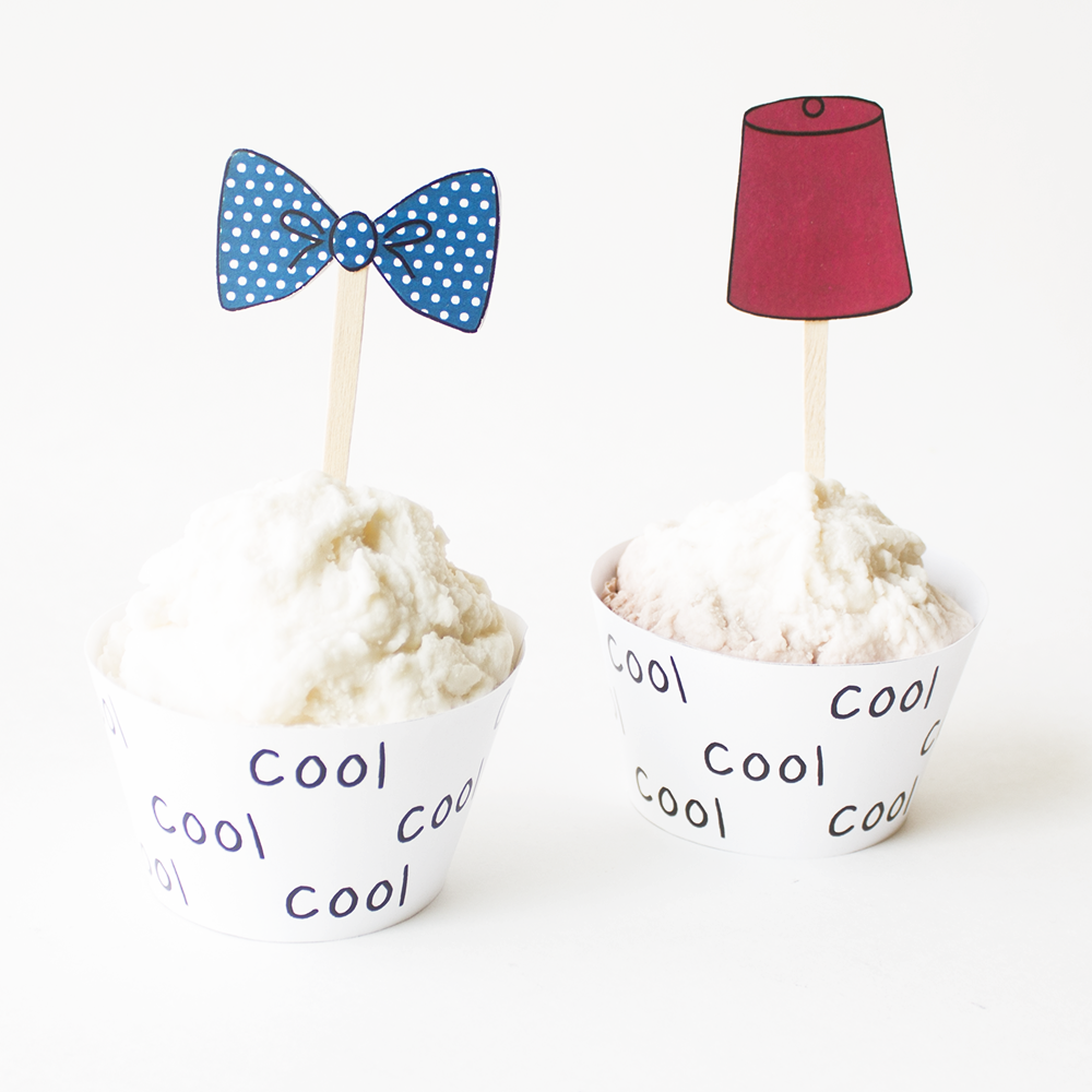 Two vanilla cupcakes on a white background. The each have a white wrapper with the word "cool" in an allover pattern. The left cupcake has a blue polkadot bow tie topper, the right has a red fez topper.