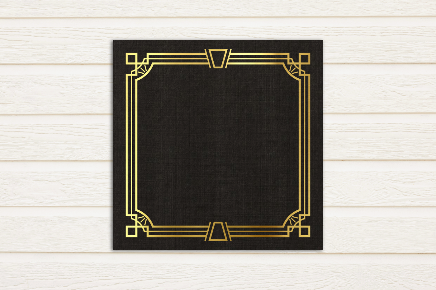 Gold art deco inspired square border design on a black square piece of paper. The paper sits on a white painted wood background.