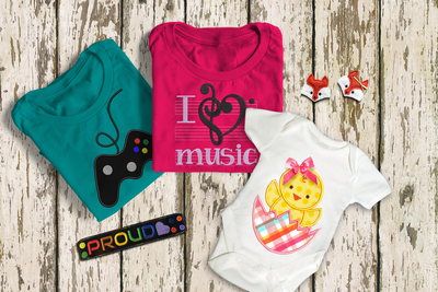 2 embroidered tees & a onesie on a wood background. At the top right are 2 fox face felties, at the lower left is a PROUD bracelet. The left shirt has a game controller, the middle shirt says I love music with symbols, & the onesie has a chick in an egg