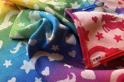 Closeup of rainbow gradient woven fabric with white silhouettes of stars, hearts, rainbows, and unicorns. The reverse is white with rainbow symbols. Tag has 3 rainbow gradient stars.