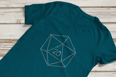 A dark teal shirt lays against a wood background. On the shirt is a d20 Die design made from clear rhinestones. The number 20 is in the center of the die.