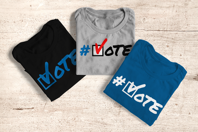 Three folded shirts against a wood background. All have the word "VOTE" with a square with a checkmark in place of the O. One is black with a blue design, one is blue with a white design, and one is grey with red, black, white and blue.