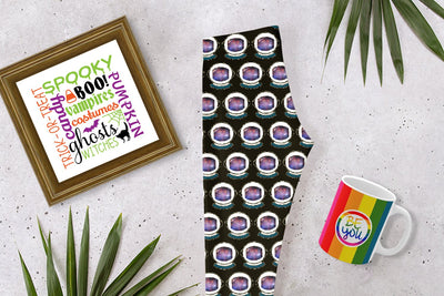 Laying on a cement surface are a framed square poster with Halloween themed words and images, a black pair of leggings with an astronaut helmet space pattern, and a mug with rainbow stripes and a circle in the center that says "Be You."