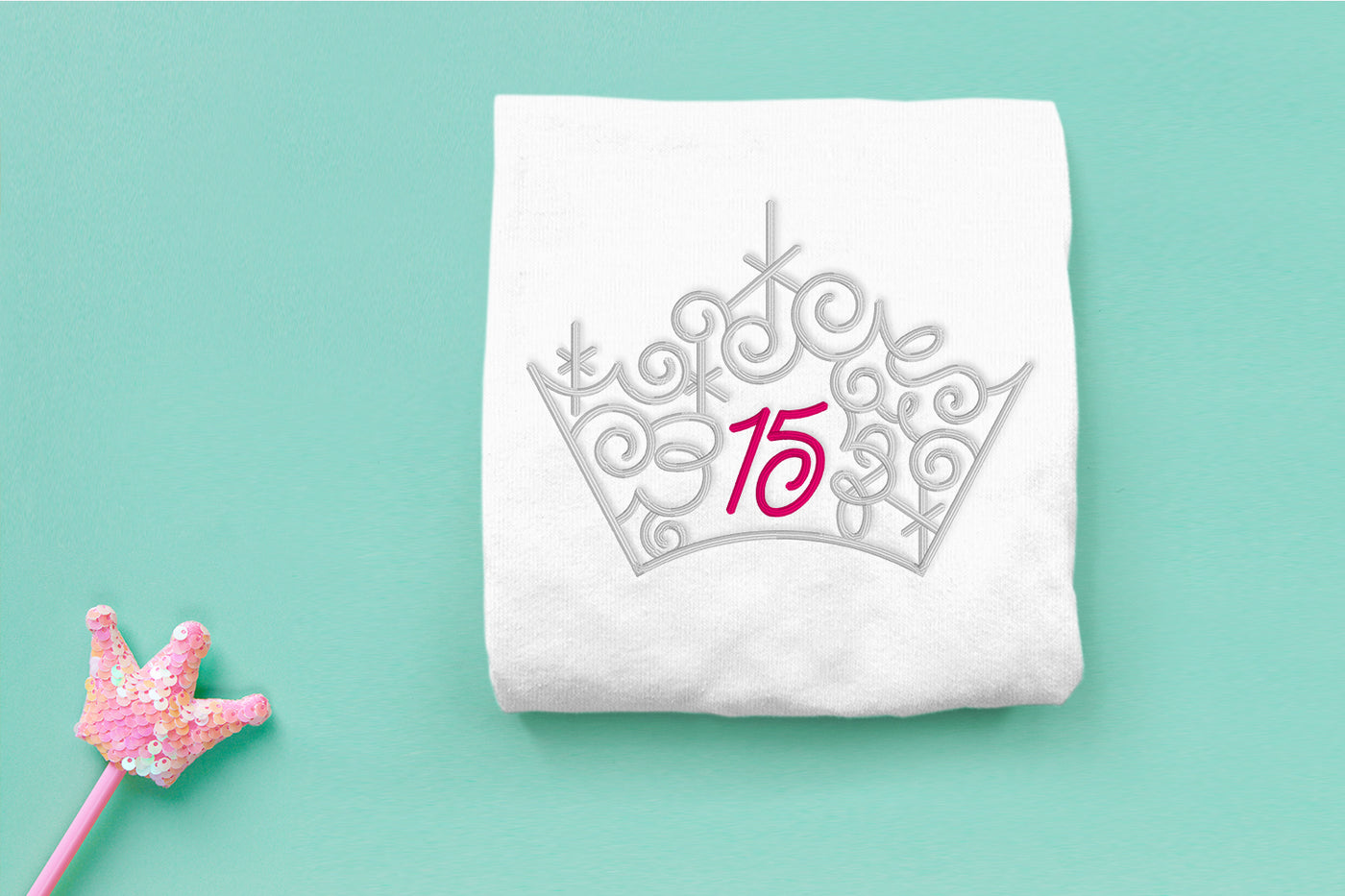 White shirt with a silver tiara design featuring the number 15 on a light aqua background.