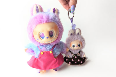 Two Labubu plush keychains, one in a pink bib dress and the other in a brown polkadot bib dress. Shows the 17 cm and 8 cm sizes of monster plush.