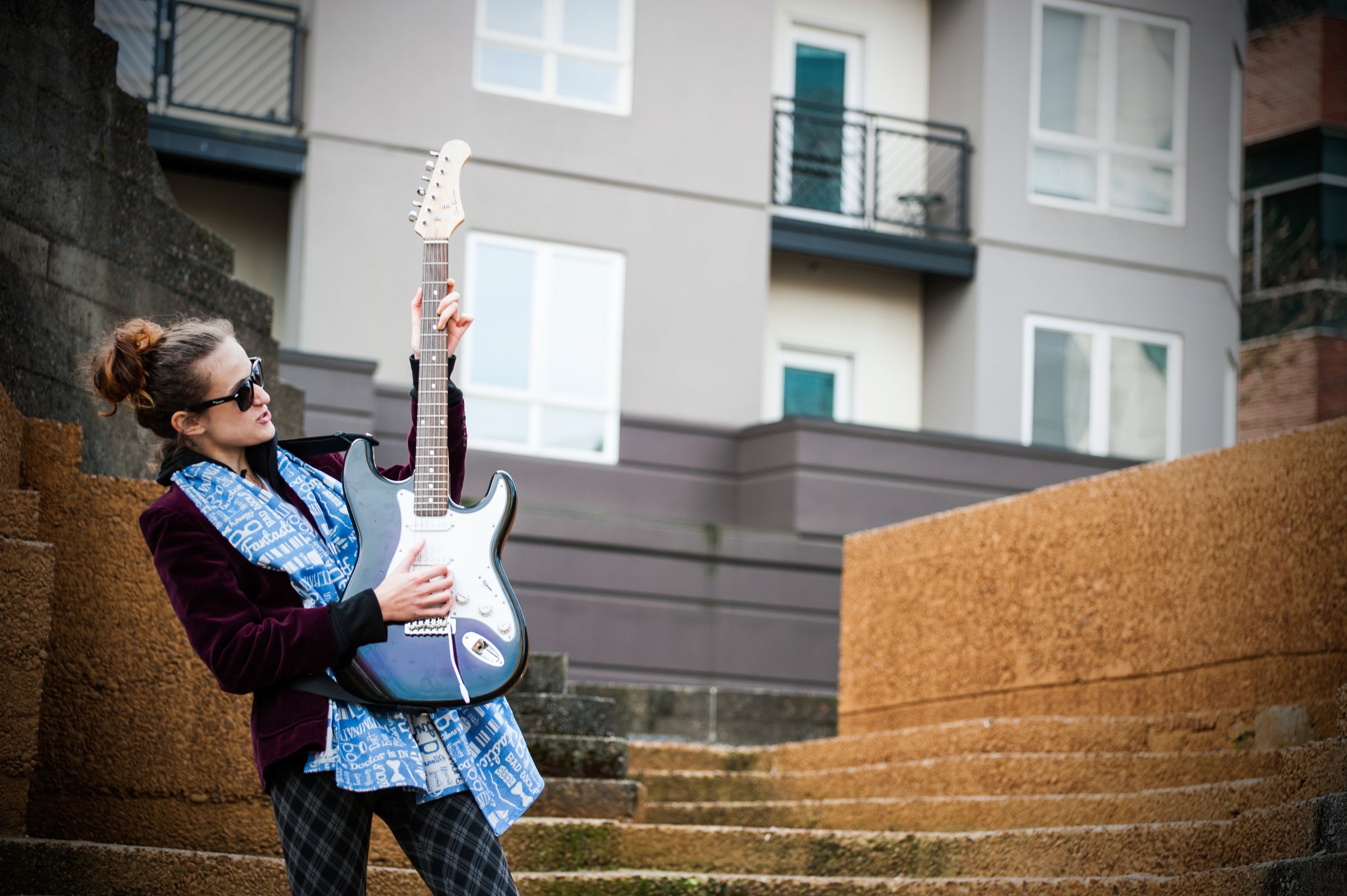 White-presenting teen girl cosplayed as the 12th Doctor from Doctor Who wears a blue and white woven fabric scarf while playing an electric guitar in front of an urban building.