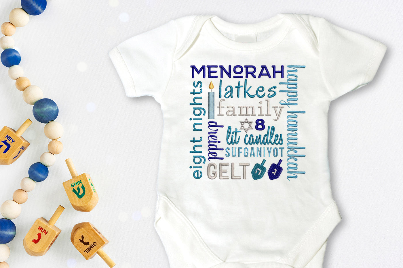 White baby onesie with Hanukkah-themed text and images embroidered onto it.