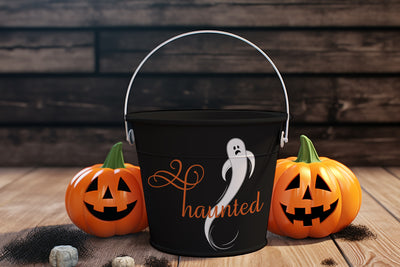 Black Halloween bucket with ghost design and 'haunted' text, surrounded by carved pumpkins on a wooden surface.