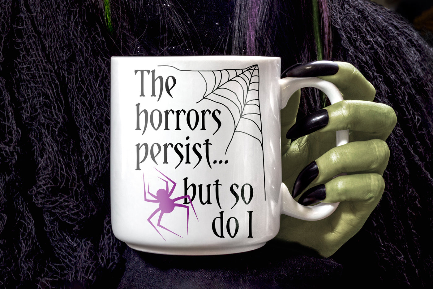 White mug with spider design and text "The horrors persist... but so do I" held by a witch hand with black nails 