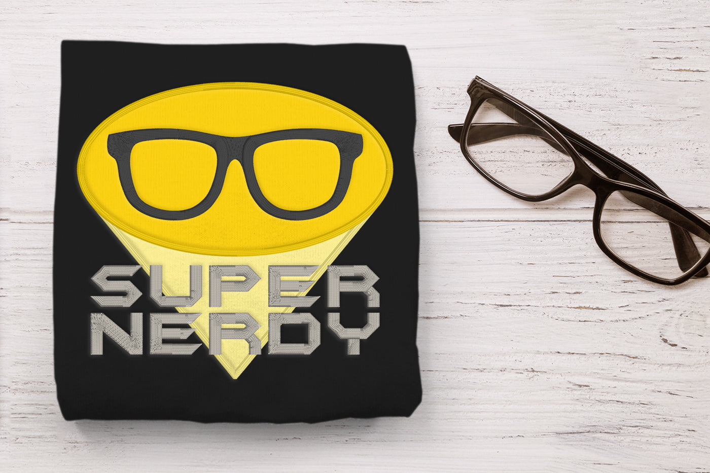 Black t-shirt with applique yellow superhero logo with glasses and 'Super Nerdy' text on a white wooden surface with glasses.