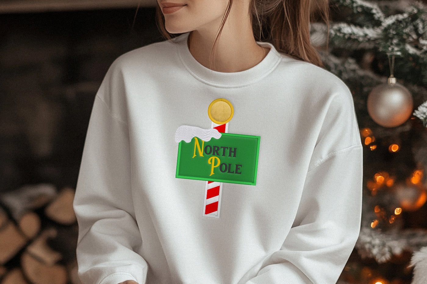 Person wearing a white sweatshirt with a 'North Pole' sign applique design in front of a Christmas tree.