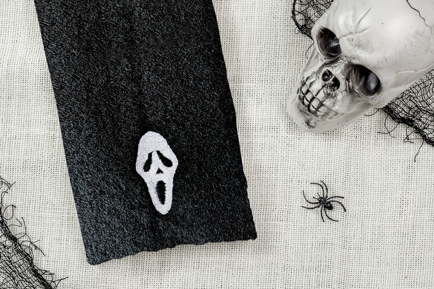 Black towel embroidery with white ghost face design next to a skull and spider on a textured surface