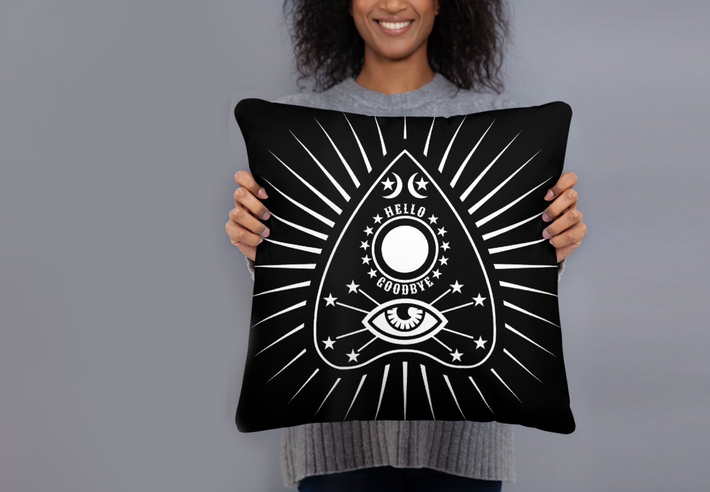 A smiling black woman holds a throw pillow in front of her. The pillow is black with a white communication board planchette design with lines radiating out from it. She stands against a grey wall.