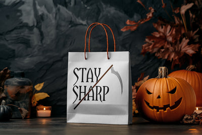 Halloween-themed paper bag with 'Stay Sharp' text and scythe. Bag is surrounded by Halloween pumpkins and  decorations.