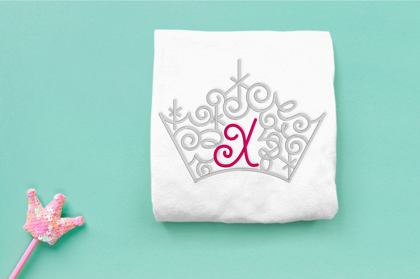 White shirt with a silver crown design and pink letter 'X' on a teal background, with a pink princess wand.