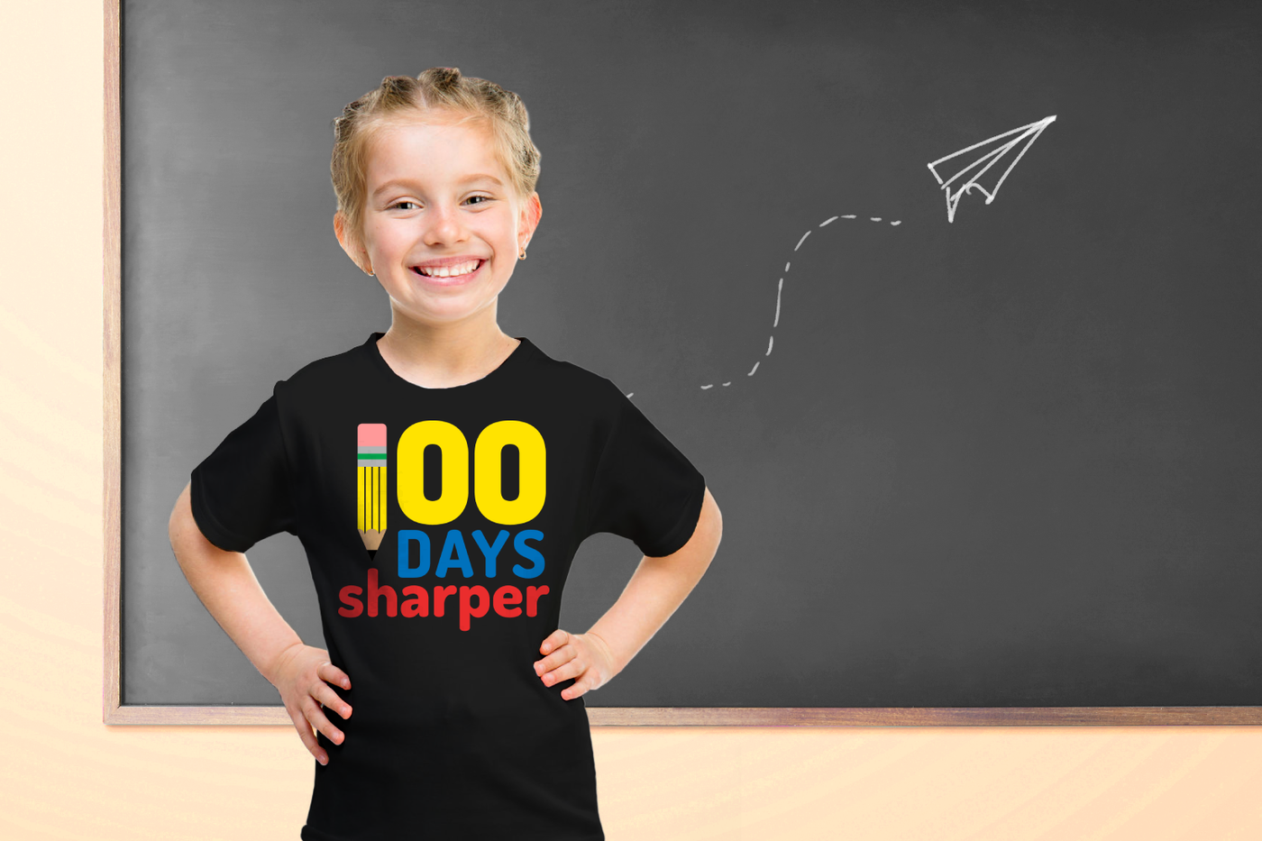 A white little girl wearing a black tee stands in front of a chalkboard with a paper airplane drawn on it. Her shirt says "100 days sharper" in yellow, blue, and red with a pencil in place of the 1.