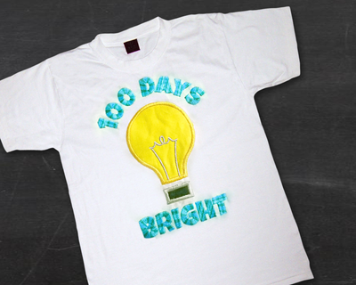 Applique light bulb on a white shirt with embroidered text in blue that says "100 days bright"