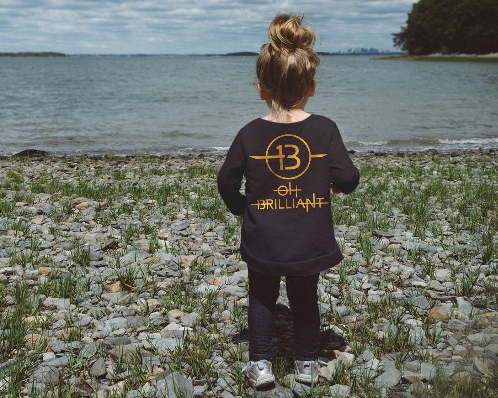 A white toddler is seen from behind, standing at the edge of a lake. She wears a black shirt with a 13 in a circle and the words "oh brilliant" below.