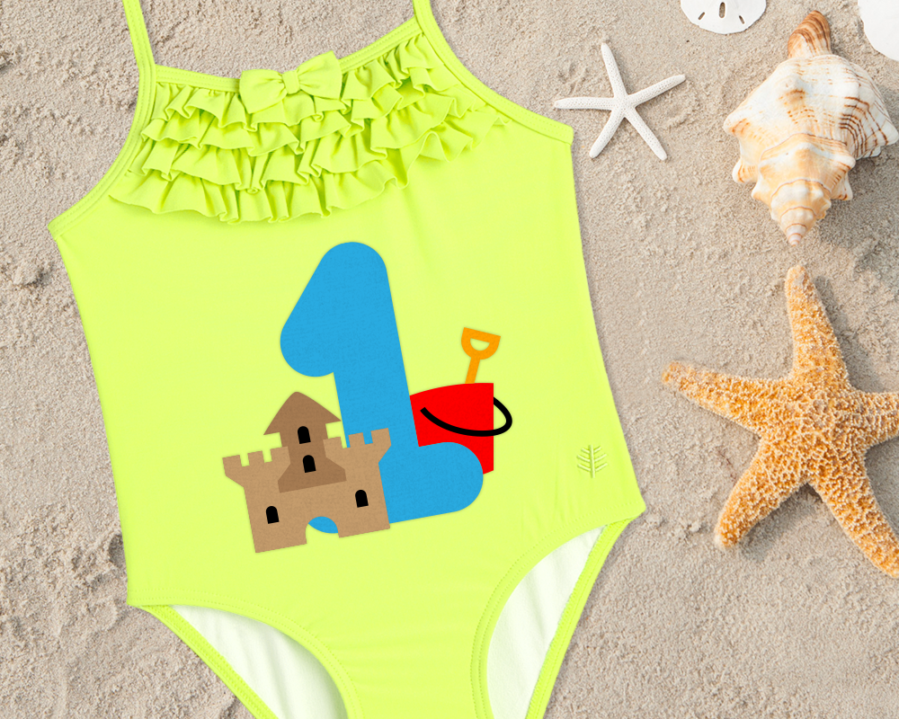Baby swimsuit with a large 1 on it. In front of the number is a sand castle, and behind it is a red pail.