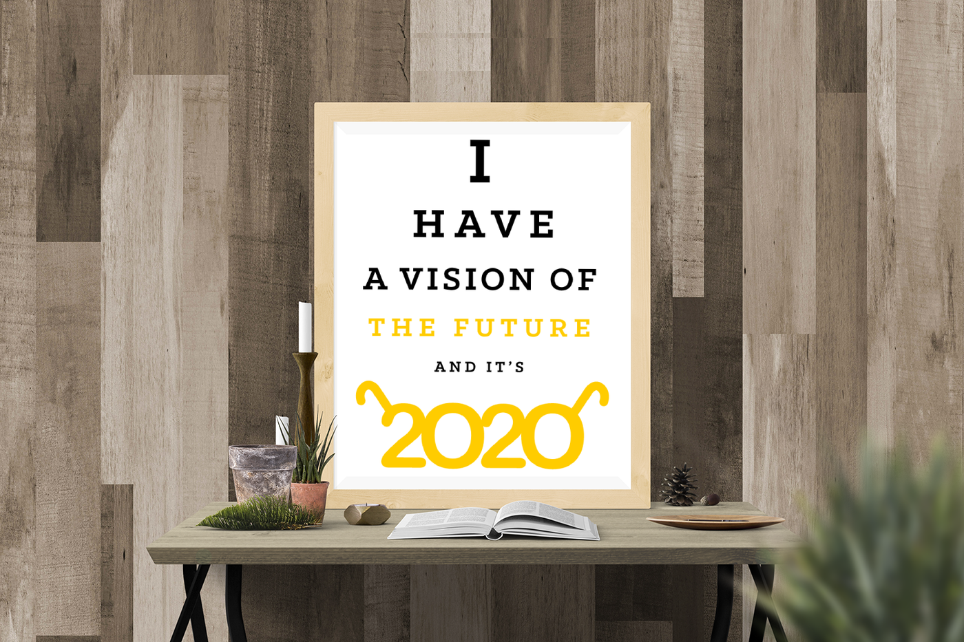 A framed poster sits on a desk against a wood panel wall. The poster is white with a yellow and black design that reads "I have a vision of the future and it's 2020." The words resemble an eye chart. The 2020 has arms to look like a pair of glasses.