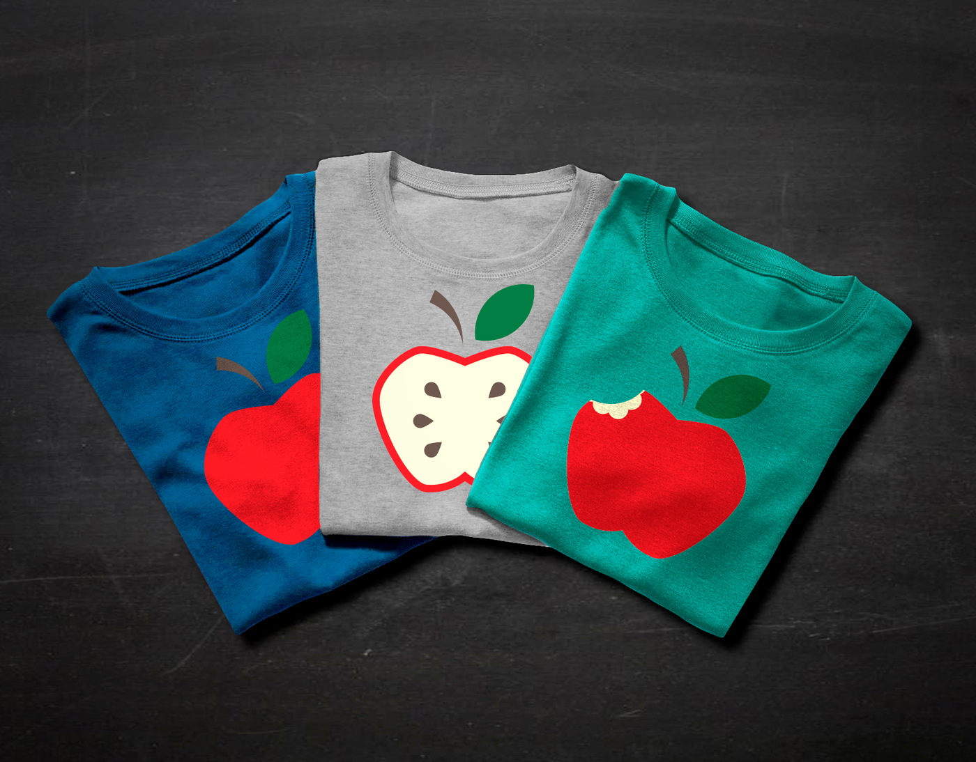 Three folded tees fanned out against a chalkboard backdrop. The left has a whole apple design, the middle has an apple that has been sliced in half to show the seeds, and the right has a design of an apple with a bite taken out.