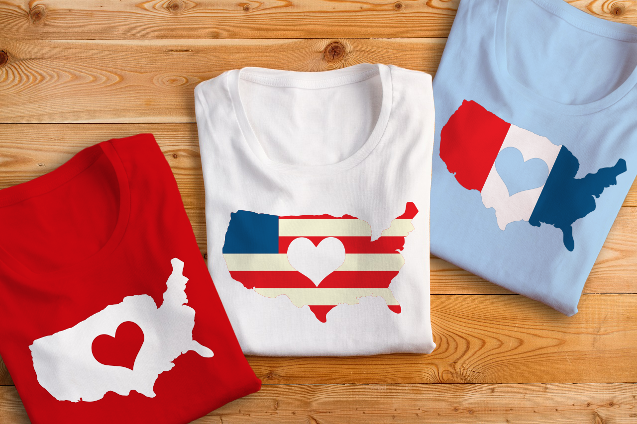 Three folded shirts, each with an image of the United States. One is a single color with a heart knockout, one has stripes like the American flag and a heart knockout, and one has vertical tri-colored bands with a heart knockout.