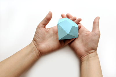 Two outstretched hands hold a 3D paper gemstone in aqua against a pale background.
