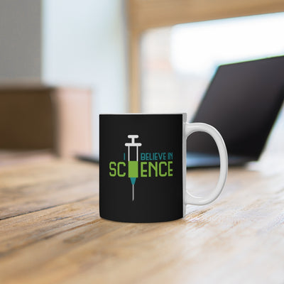 Mug with a design that says "I believe in science" with a syringe. Mug is on a desk.