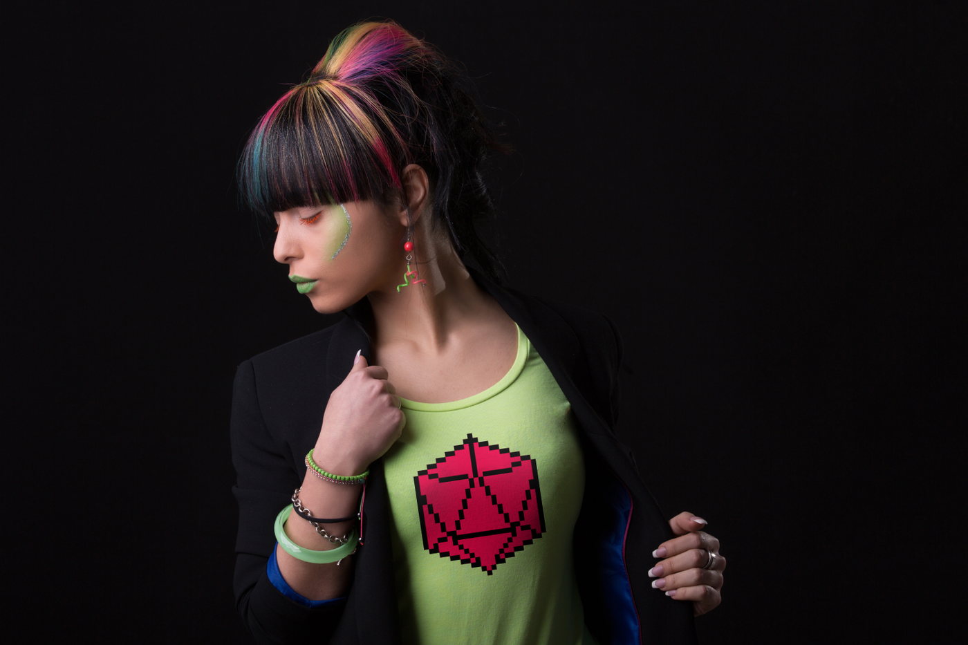A woman of color with brightly colored neon makeup and neon streaks in her hair wears a black blazer and bright green tee. On the tee is a pink D20 done in 8-bit style.
