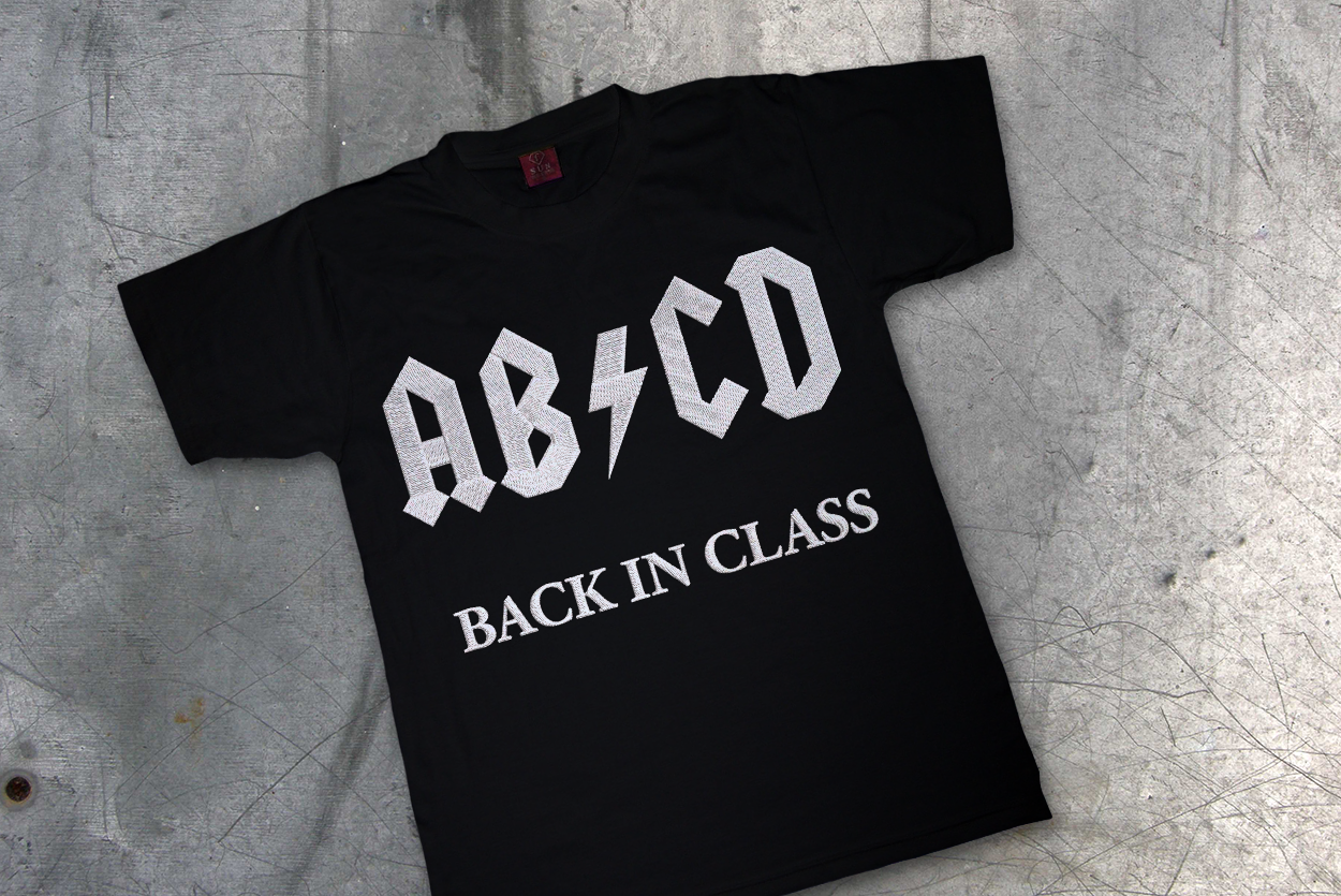 A black child's shirt on a metal background. There is a white embroidered design that says "AB/CD back in class." The slash looks like a lightning bolt and is a parody of the AC/DC logo.