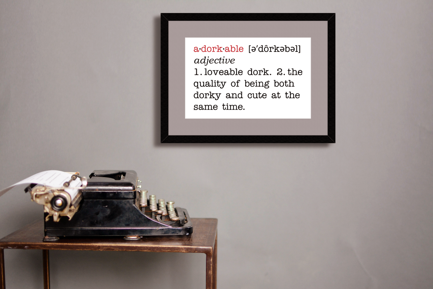 A framed poster on a grey wall above a table with a typewriter. The poster looks like a dictionary definition and says "adorkable. adjective. 1. loveable dork. 2. the quality of being both dorky and cute at the same time."