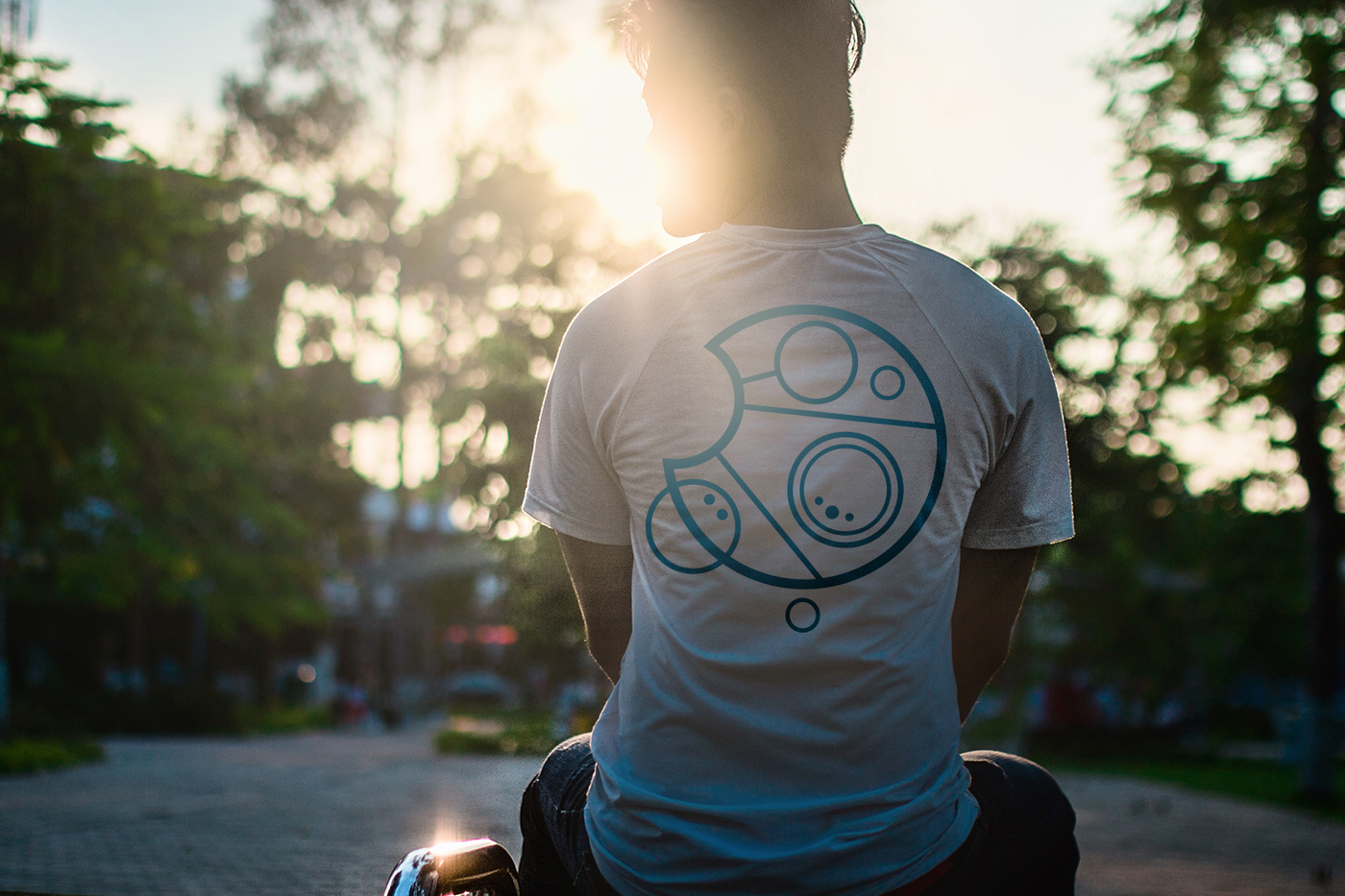 What man shown from behind sitting, backlit by the sun. He wears a white tee with a blue design in circular Gallifreyan.
