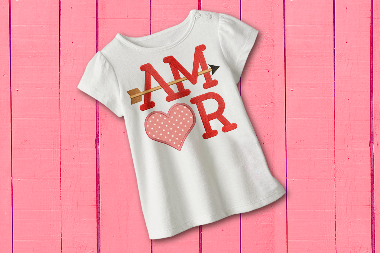 A white child's shirt lays on a pink painted wood background. Embroidered onto the shirt is the word "AMOR" set on a square. An arrow flies through the A and M. The O is an applique heart with pink polkadot fabric.
