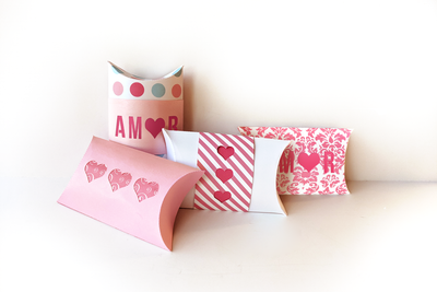 Four pillow boxes sit on a white background. They are done in shades of pink and white. Two have cutouts of 3 hearts, revealing a second paper pattern, the other two have "AMOR" cut out with a heart for the O.  Two have decorative bands around them.