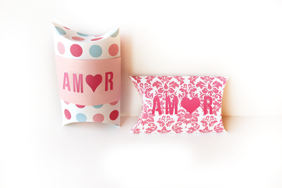 Two pillow boxes on a white background. On box is white with pink and blue dots and a pale pink band with the word "AMOR" cut out to reveal dark pink behind it. The other box is pink damask on white with the word "AMOR" cut out to reveal pink behind it. Both words have a heart in place of the O.