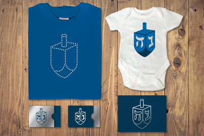 A shirt, baby onesie, card, and two gift card holders. Each has a dreidel design on it. The card is done in silver foil as a line drawing. The shirt is done out of clear rhinestones.
