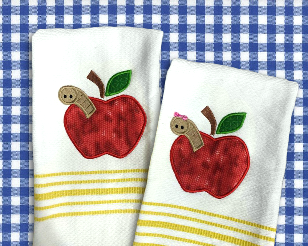 Two white and yellow tea towels lay against a blue gingham surface. Embroidered onto each towel is an apple with a work peeking out. The right worm has a little pink bow on its head.