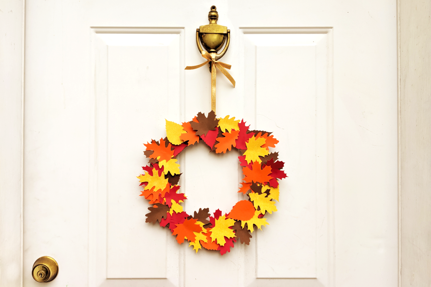 A wreath made of paper leaves in fall colors hangs on a white door with a brass knocker from a gold ribbon.