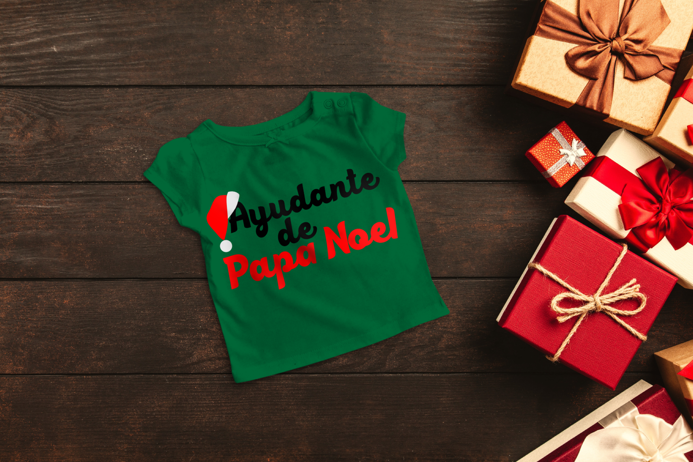A green child's shirt lays on a dark wood background with red and gold wrapped gifts to the side. The shirt has the words "Ayudante de Papa Noel" with a Santa hat hanging off of the first A.