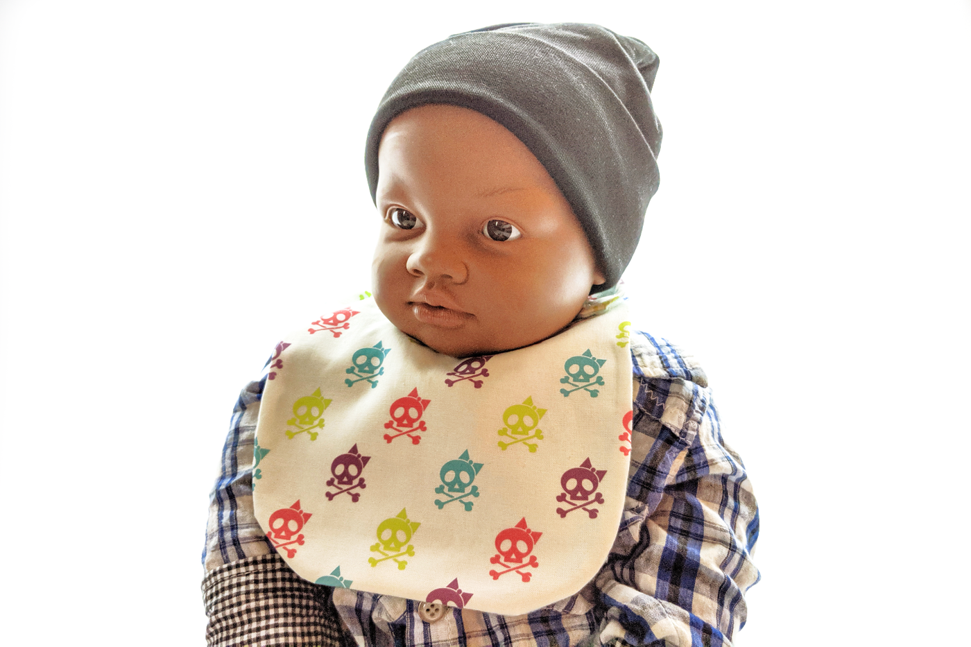 A black baby mannequin wears a bib, plaid shirt, and grey beanie. The bib fabric is off-white with a pattern of cute skulls with bows in lime green, turquoise, hot pink, and purple.