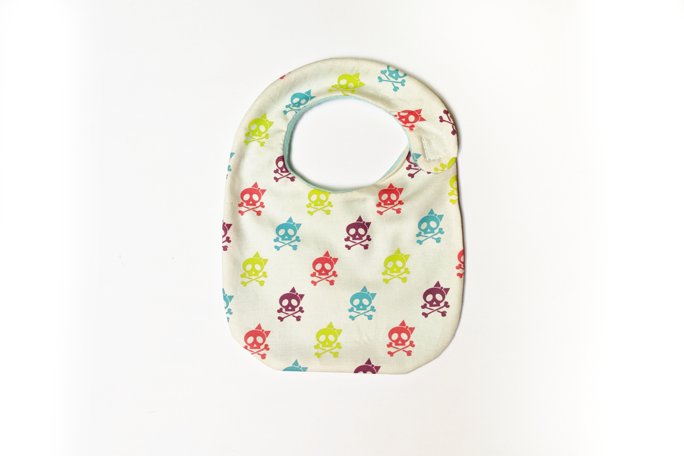 A baby bib sits on a white surface. The fabric is off-white with a pattern of cute skulls with bows in lime green, turquoise, hot pink, and purple.
