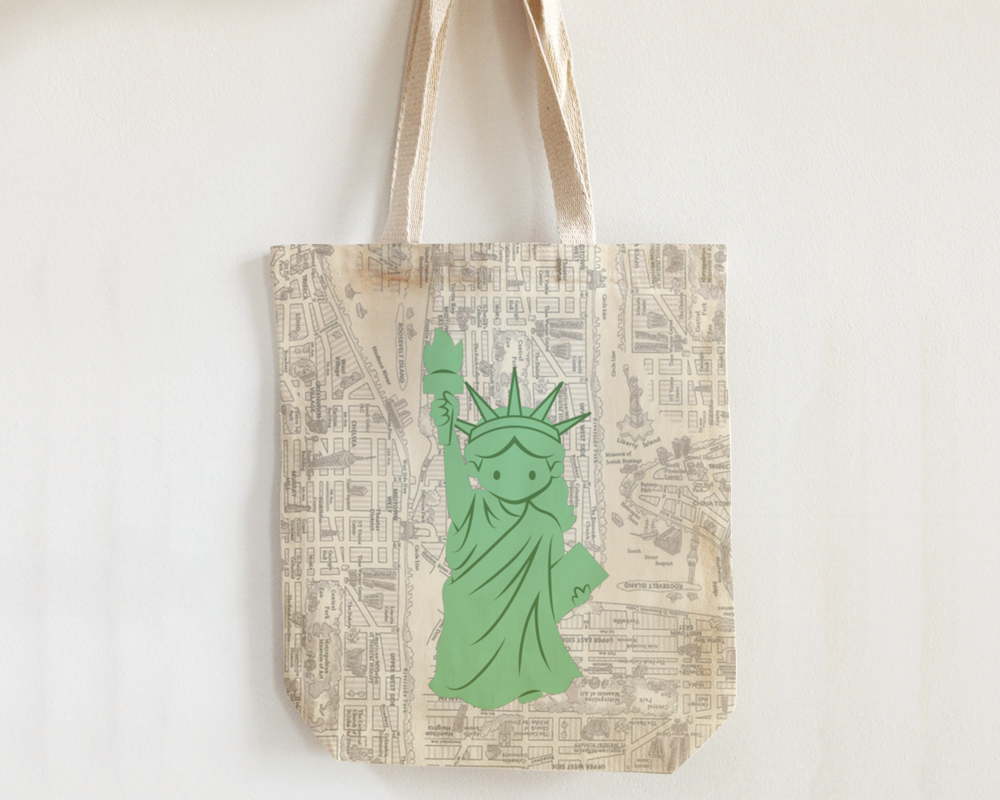 A tote bag made out of new york city map illustration fabric hangs on a white wall.  Added to the bag is a pale green Statue of Liberty illustration where she looks like a young child.
