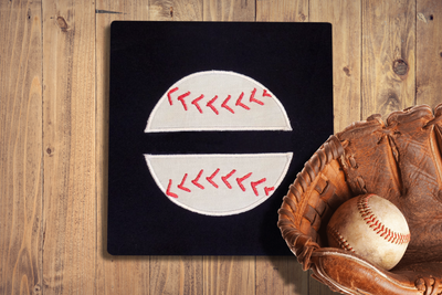 A black square of fabric sits on a wooden surface with a baseball glove holding a ball on top. Embroidered onto the fabric is a baseball with red stitching and a split space in the middle.