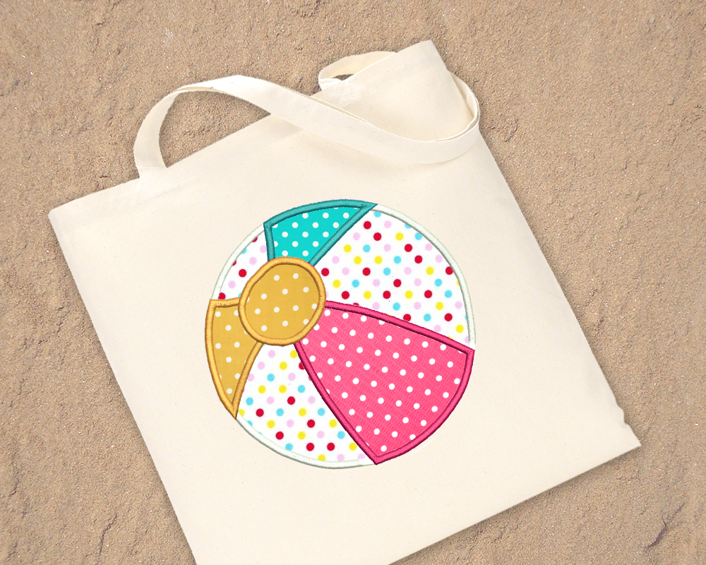 Natural colored tote bag laying on sand. Embroidered on the bag is an applique beach ball in white, pink, turquoise, and yellow polka dot fabrics.