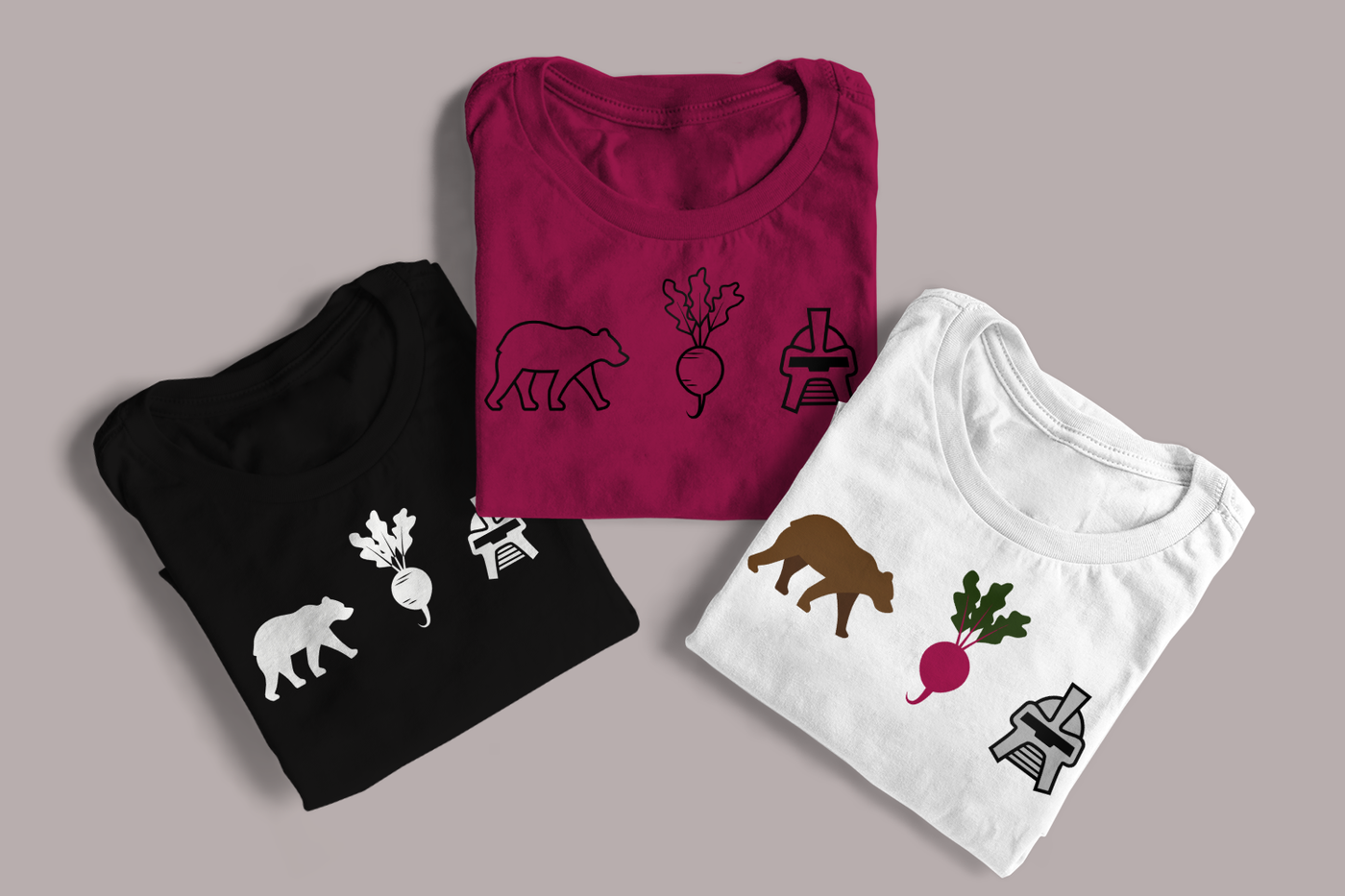 Three folded shirts on a grey background. Each shirt has the image of a bear, a beet, and a cylon helmet.