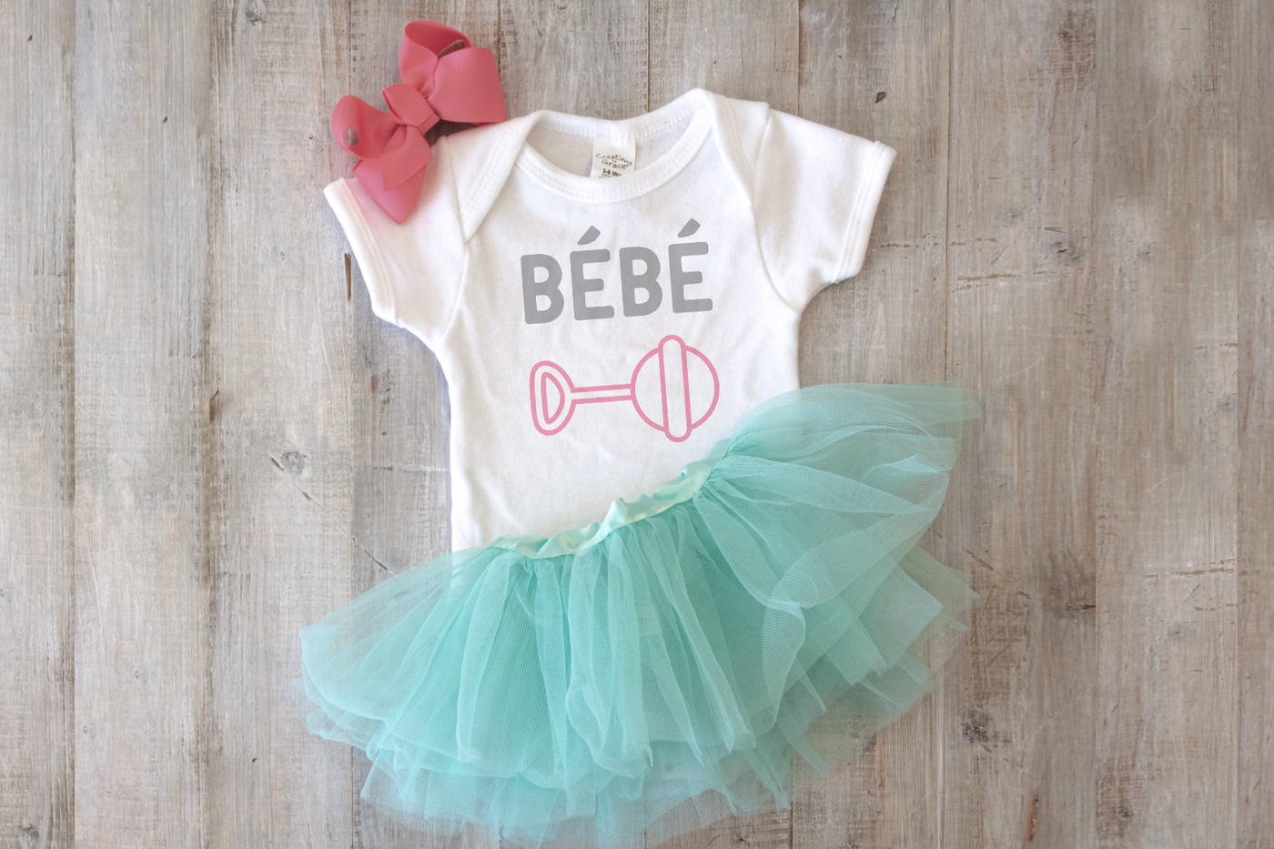 A white onesie with an aqua tutu and pink bow lays on a wooden surface. The onesie says "bébé" and has a line drawing of a rattle below.
